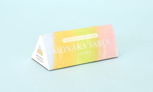 モナカサブレ用の三角柱パッケージの印刷紙器事例。特殊な三角形の形状、水彩画のようなデザインと和紙風の風合い（DKホワイトF、マットニス）、店頭で目を引くアイキャッチ効果、FSC認証マークの印刷、およびマット系特殊紙に合わせた印刷色調整の設計ポイントを示した画像。